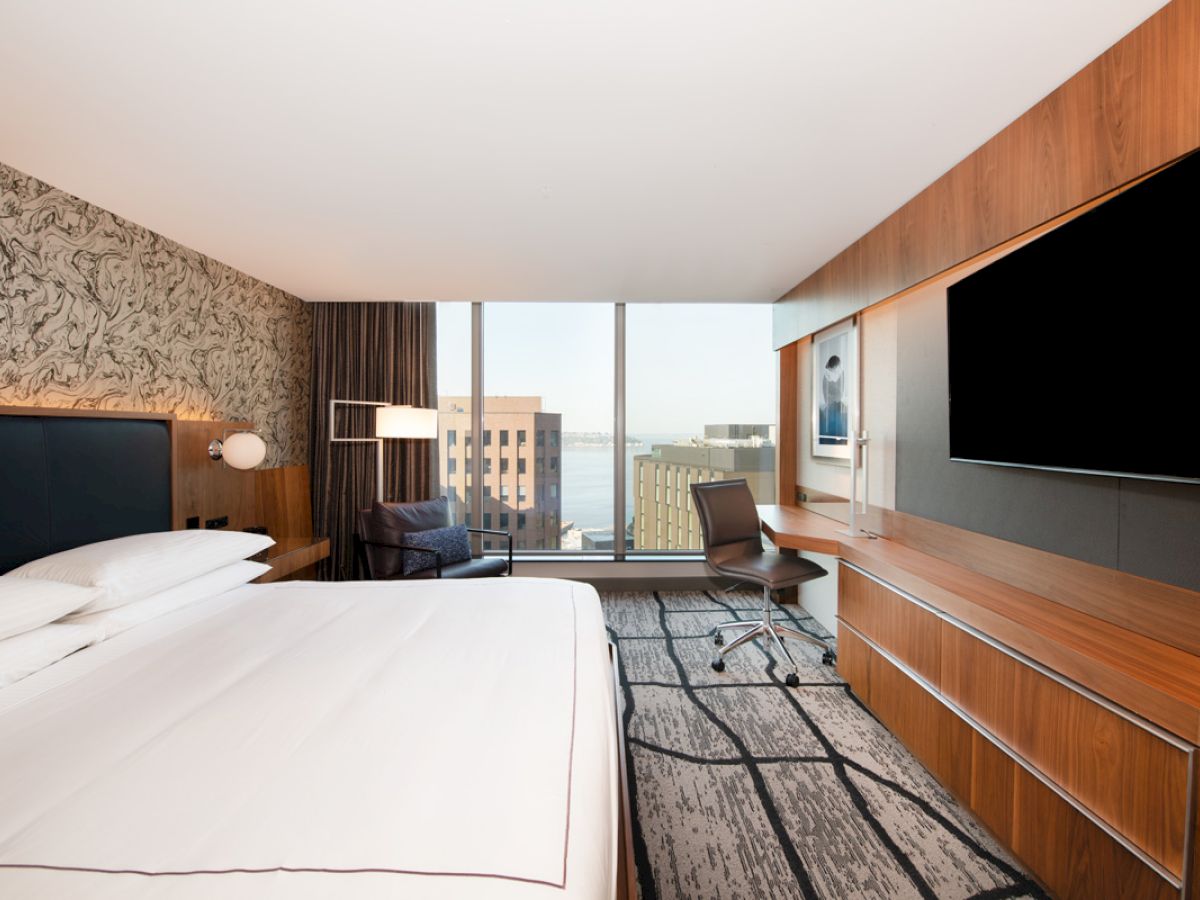 A modern hotel room featuring a large bed, wall-mounted TV, desk, chair, and a window offering a city view with buildings and water visible.