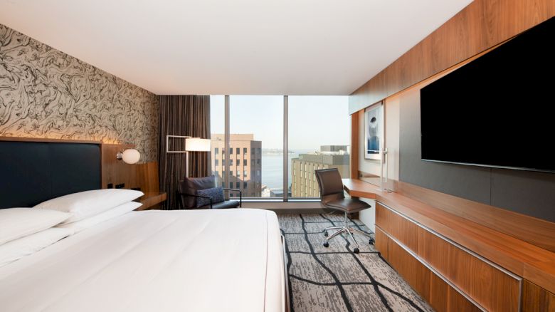 A modern hotel room featuring a large bed, wall-mounted TV, desk, chair, and a window offering a city view with buildings and water visible.