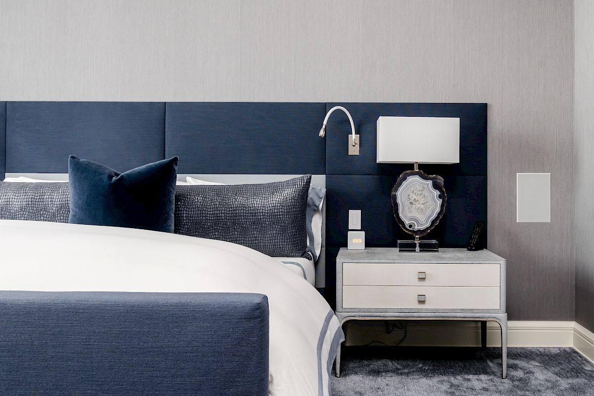A modern bedroom featuring a dark blue upholstered headboard, white bedding, side table with two drawers, a lamp, and decor.