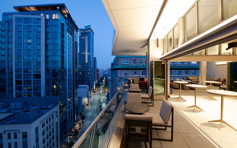 A modern urban balcony with seating and tables overlooks a street lined with tall buildings at dusk, showcasing city lights and architecture.