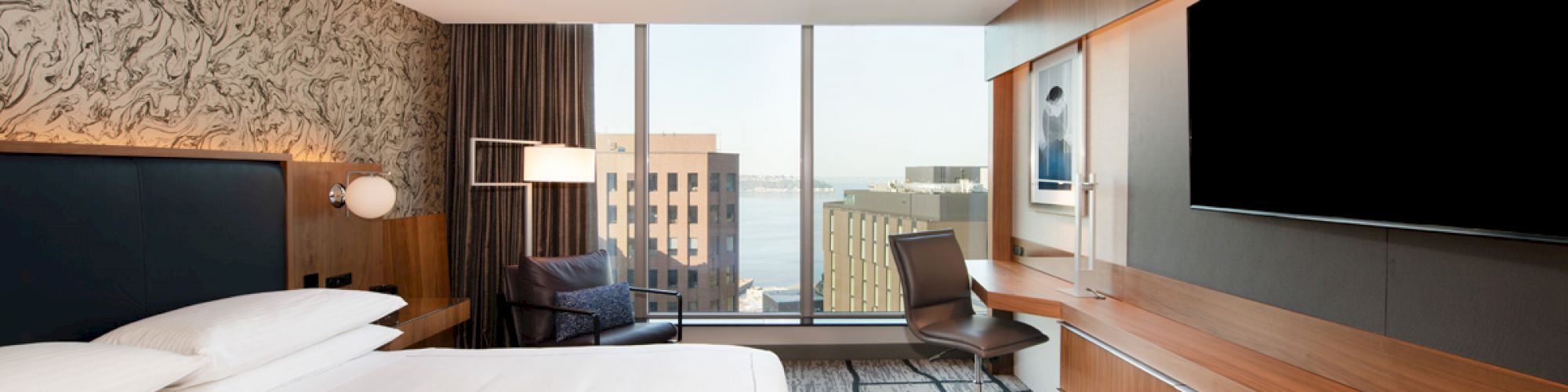 This image shows a modern hotel room with a large bed, a flat-screen TV, a desk, and a view of buildings through a large window.