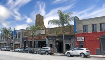 A street scene with palm trees, various parked cars, and buildings including a theater and a hotel against a backdrop of blue sky.