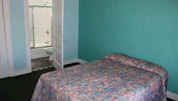 A small bedroom with a bed covered in a colorful quilt and an open door leading to a bathroom with a shower and toilet.