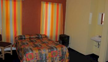 A small bedroom with a double bed, colorful bedspread, striped curtains, a chair, a wall-mounted sink, and a small mirror.