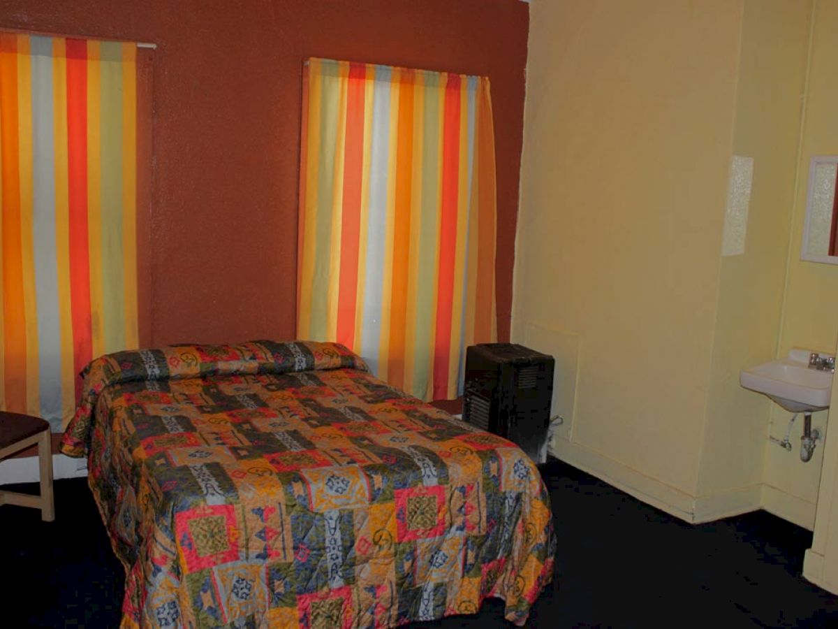 A small hotel room with a single bed, colorful curtains, a chair, a small nightstand, and a sink.