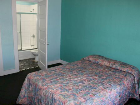 The image shows a small bedroom with a colorful bedspread, and an open door leading to a bathroom with a shower and a toilet.